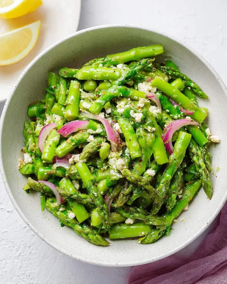 A vibrant asparagus salad in a light-colored bowl, featuring bright green asparagus, red onion slices, and crumbled feta cheese, with lemon wedges in the background.