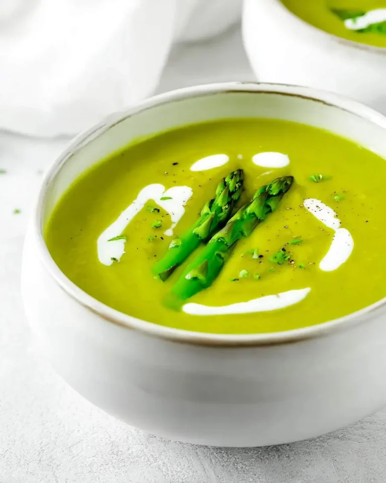 A close-up of a white bowl filled with vibrant green asparagus soup, garnished with two whole asparagus spears, cream swirls, and black pepper.