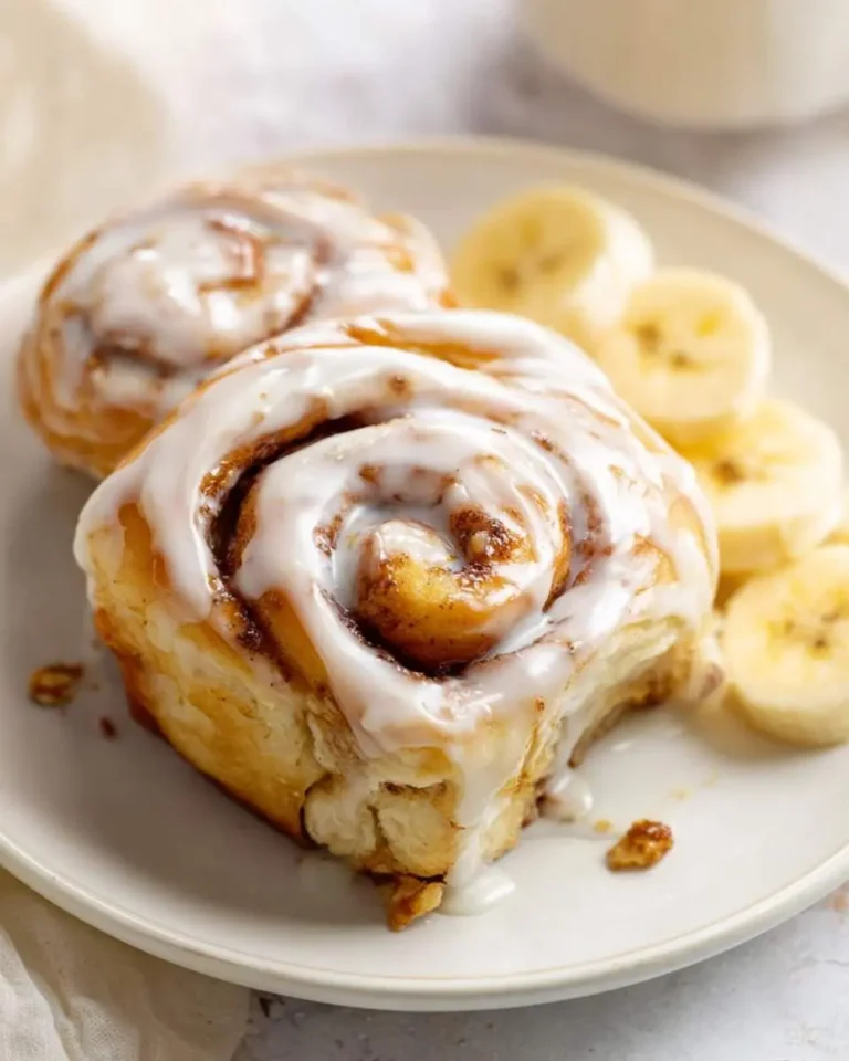 A plate with two glazed Banana Bread Cinnamon Rolls and sliced bananas, one roll has a small bite taken out.