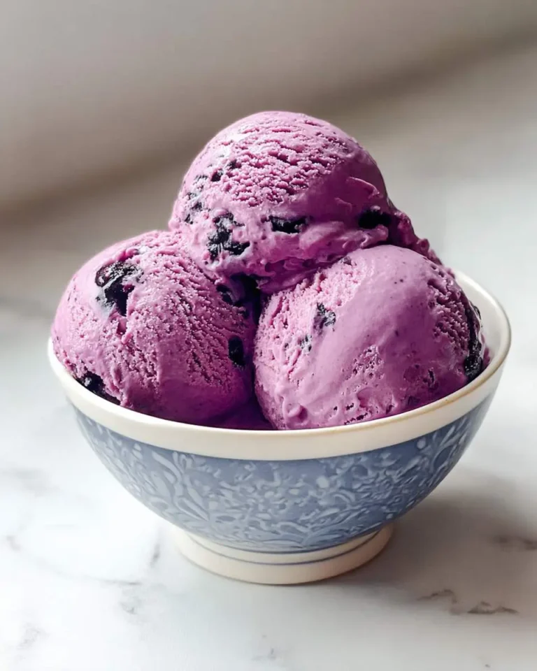 Three scoops of vibrant purple blueberry ice cream with dark fruit pieces, served in a decorative blue and white bowl.