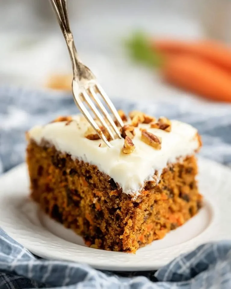 A fork is digging into a delicious slice of Carrot Cake Recipe with Cream Cheese Frosting, topped with chopped pecans, on a white plate.