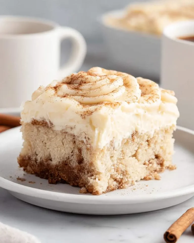 A delightful slice of cinnamon roll poke cake with creamy white frosting and a dusting of cinnamon, served on a white plate.
