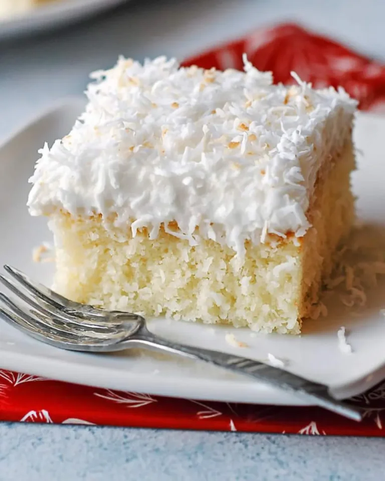 A delicious slice of Coconut Cream Cake topped with white frosting and shredded coconut, served on a white plate with a fork.