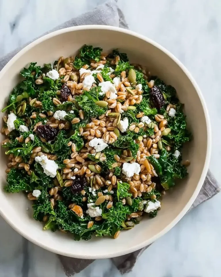 A vibrant Farro Kale Salad With Goat Cheese, tossed with plump raisins and green pumpkin seeds in a white bowl.