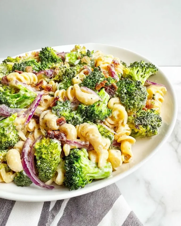 A close-up shot of a creamy and vibrant Fresh Broccoli Pasta Salad, featuring green broccoli florets, pasta, red onion, and bacon bits in a white dish.