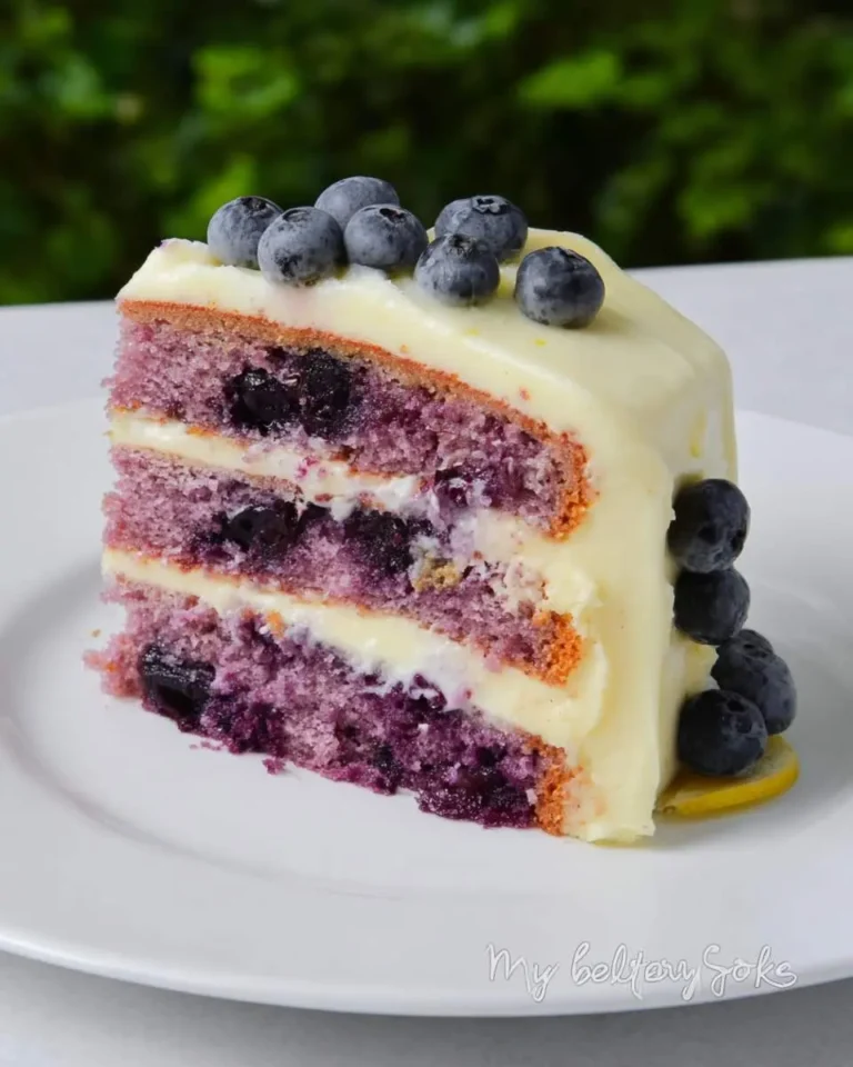 A vibrant slice of Lemon Blueberry Cake with Cream Cheese Frosting, with purple layers, baked blueberries, and fresh fruit topping.