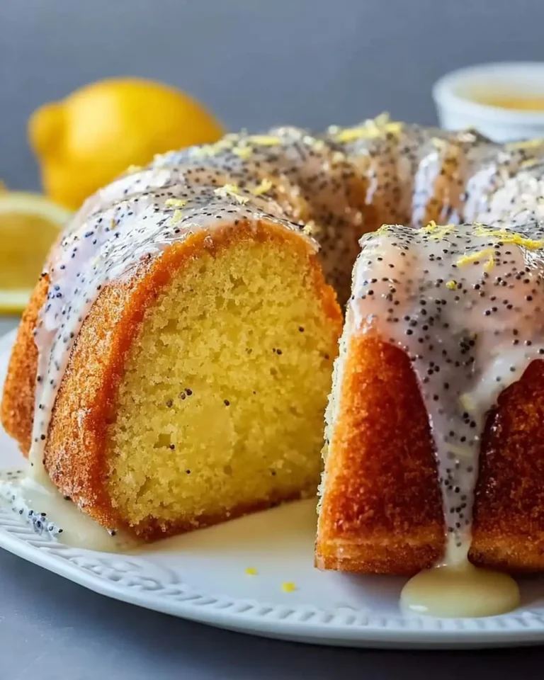 A golden Lemon Poppy Seed Bundt Cake, with a slice cut out, generously drizzled with white lemon glaze and sprinkled with lemon zest, on a white plate.