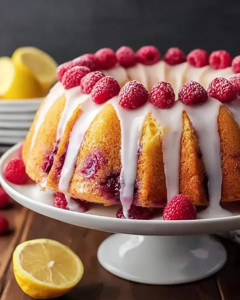 A delightful Lemon Raspberry Bundt Cake on a white cake stand, drizzled with white icing and topped with fresh raspberries, with lemons in the background.