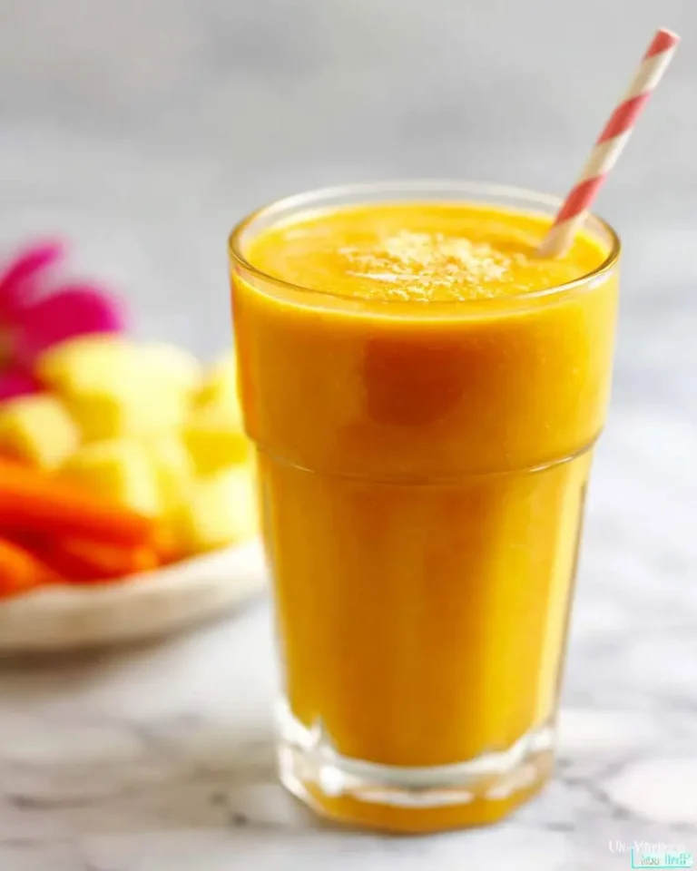 A vibrant mango carrot smoothie in a tall glass with a striped straw, garnished. Blurred fresh carrots and pineapple are behind it.