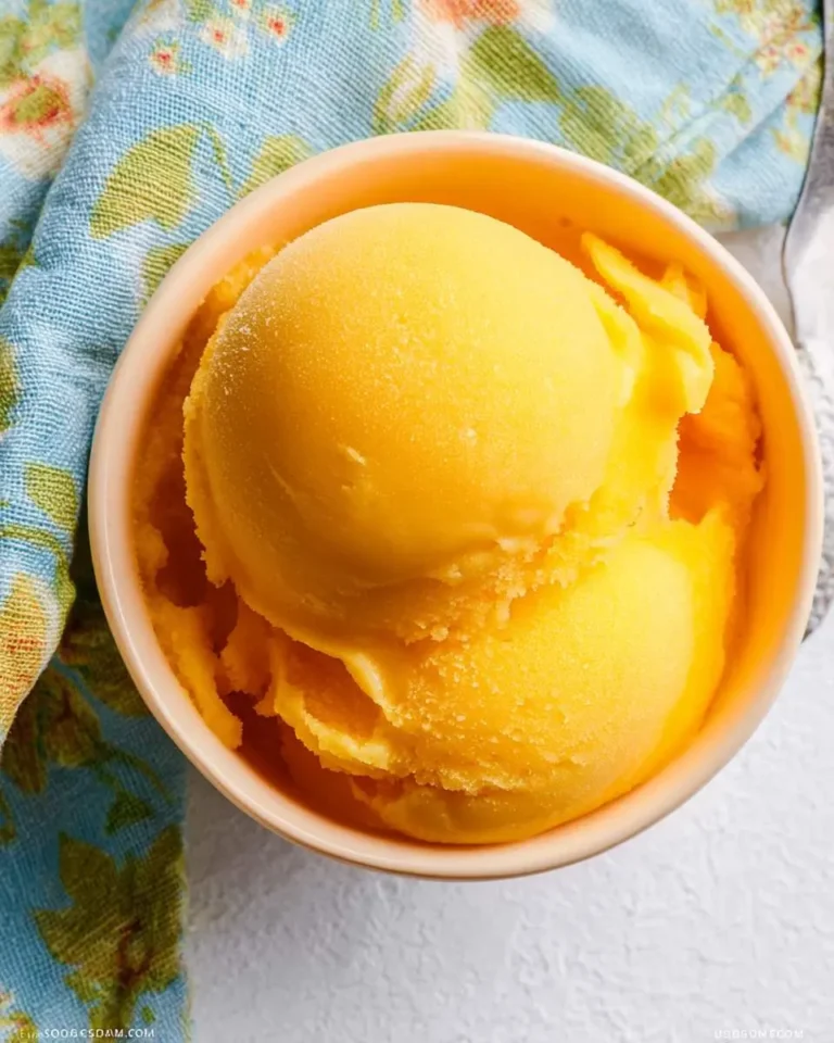 Top-down view of a bowl filled with vibrant yellow mango sorbet, placed on a white surface next to a blue floral napkin.
