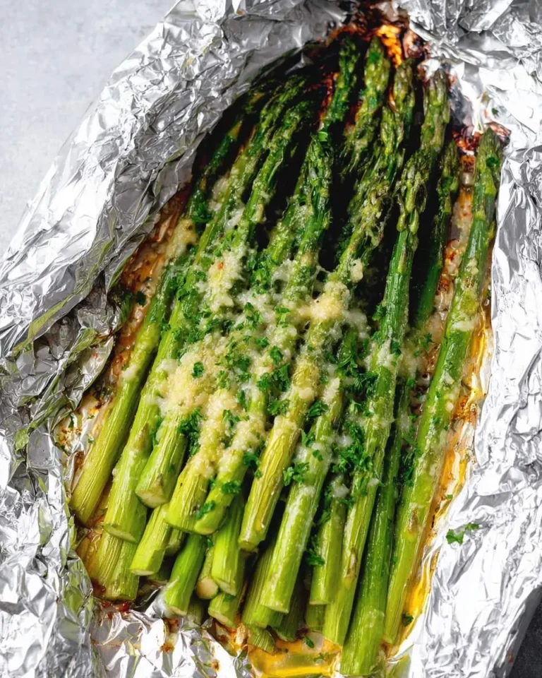 A close-up view of vibrant green Parmesan grilled asparagus in a foil packet, topped with melted cheese and fresh parsley.