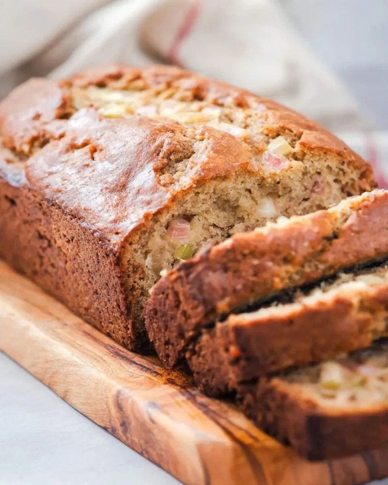 A freshly baked loaf of golden-brown rhubarb banana bread, partially sliced, resting on a rustic wooden board.