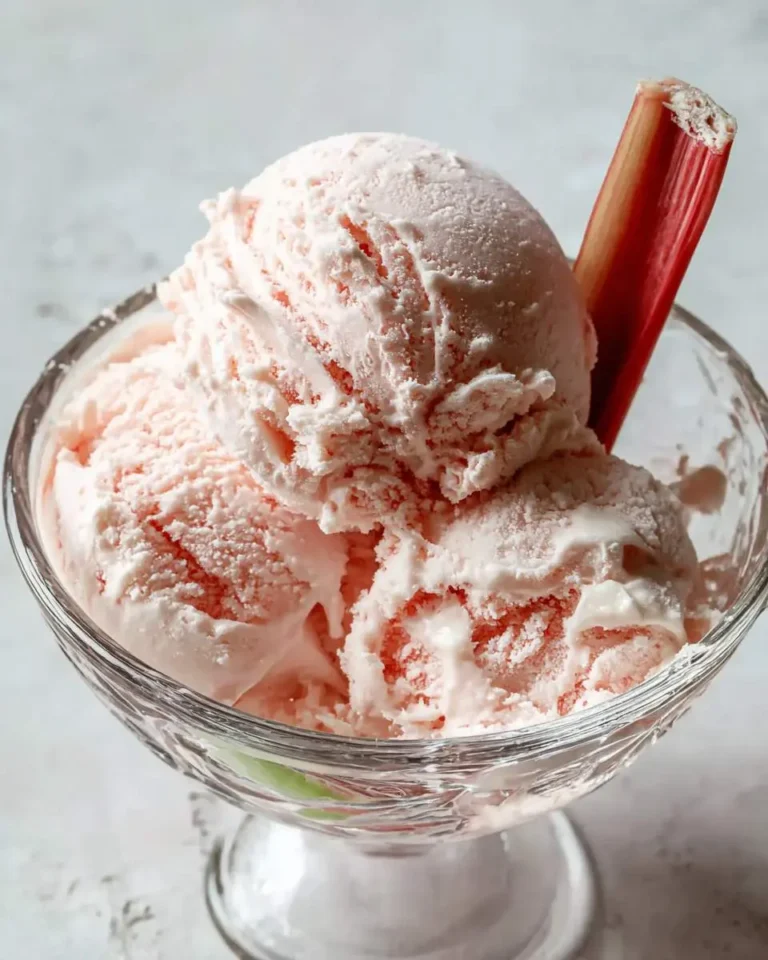 Three scoops of pink Rhubarb Ice Cream served in a clear glass dessert bowl, garnished with a fresh rhubarb stalk.