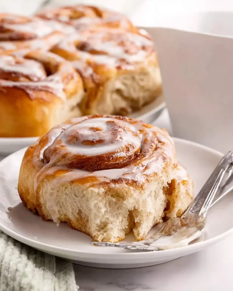 A close-up of a single, fluffy sourdough discard cinnamon roll with sweet icing, a bite taken out, and a fork on a white plate.