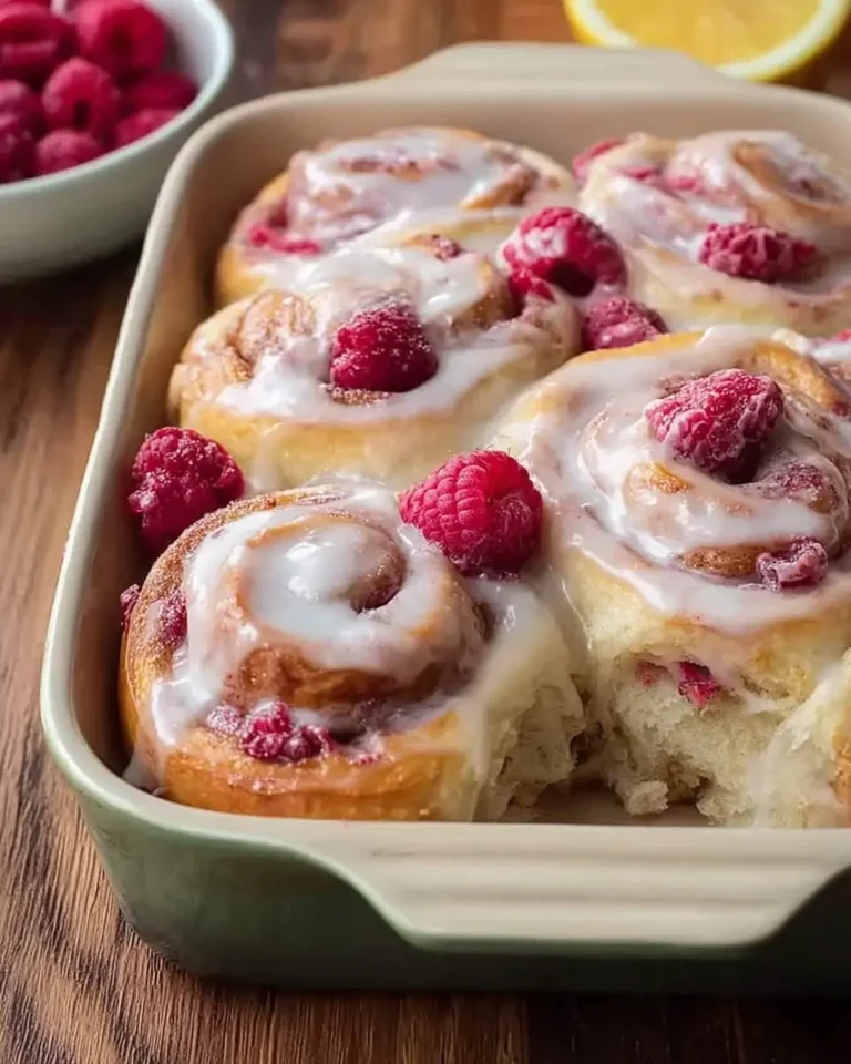 A close-up of delicious Sourdough Raspberry Lemon Rolls in a baking dish, generously topped with white icing and fresh raspberries.