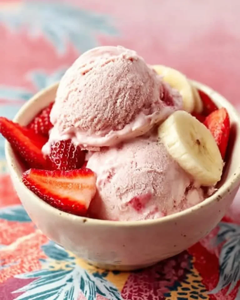 A close-up shot of creamy strawberry banana ice cream in a white bowl, garnished with fresh strawberries and banana slices.