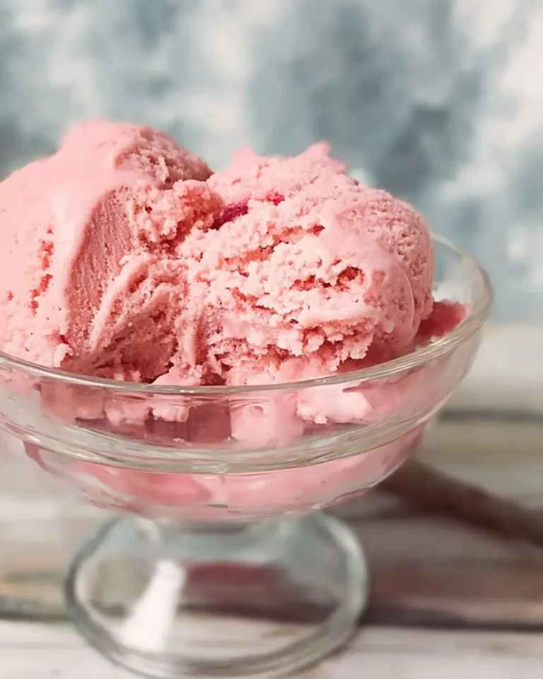 A clear glass bowl filled with several scoops of creamy pink strawberry rhubarb ice cream, slightly melting, on a light wooden surface.