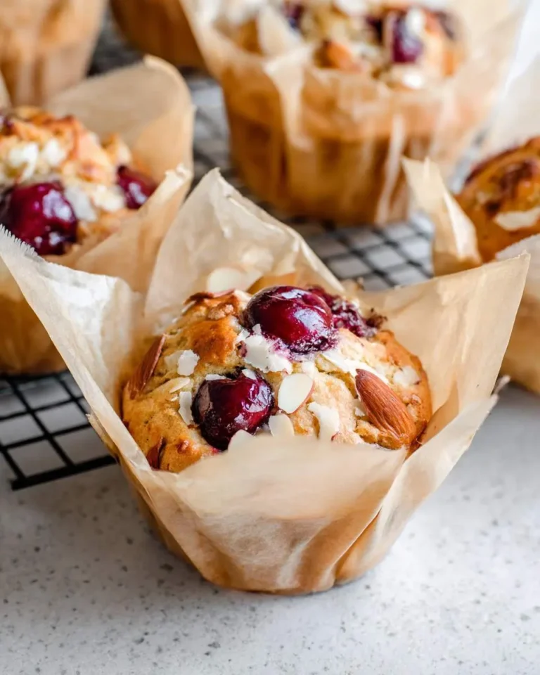 Golden brown Almond Cherry Muffins in brown paper liners, topped with glistening red cherries and crunchy almond slices, cooling on a rack.