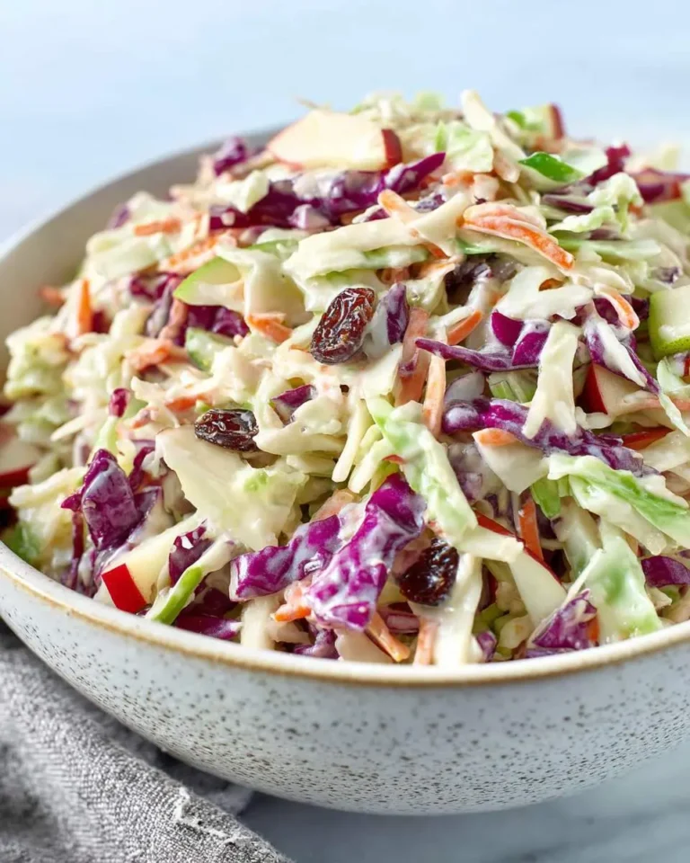 A close-up of a vibrant bowl of creamy Apple coleslaw with red cabbage, green cabbage, carrots, red and green apple pieces, and dark raisins.
