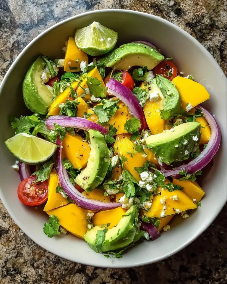 A vibrant bowl of fresh Avocado Mango Salad with red onion, cilantro, cherry tomatoes, and lime wedges.