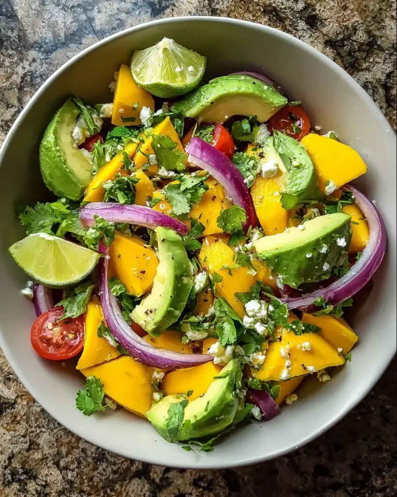 A vibrant bowl of fresh Avocado Mango Salad with red onion, cilantro, cherry tomatoes, and lime wedges.