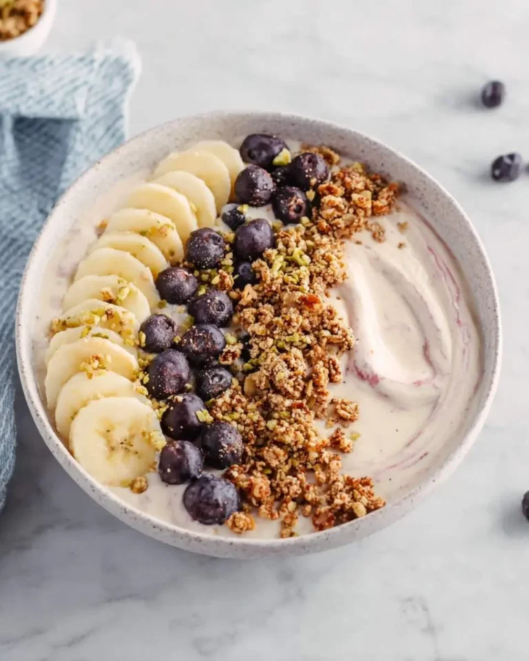 A beautiful Banana Smoothie Bowl topped with sliced bananas, fresh blueberries, crunchy granola, and chopped pistachios on a marble surface.