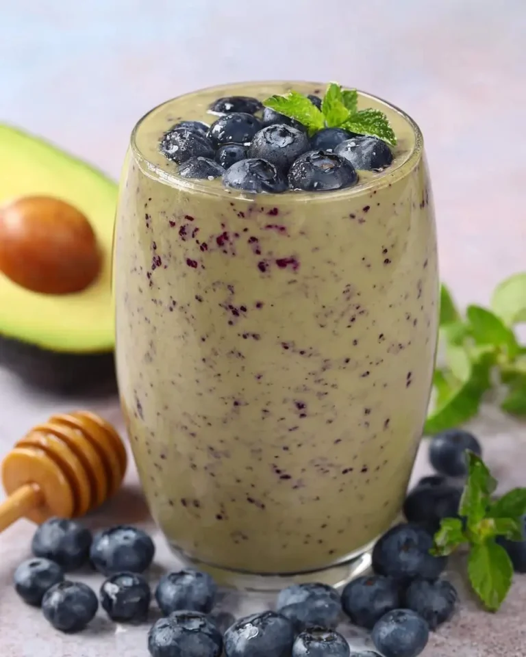 A vibrant green blueberry avocado smoothie in a glass, topped with fresh blueberries and mint, next to a halved avocado and honey dipper.