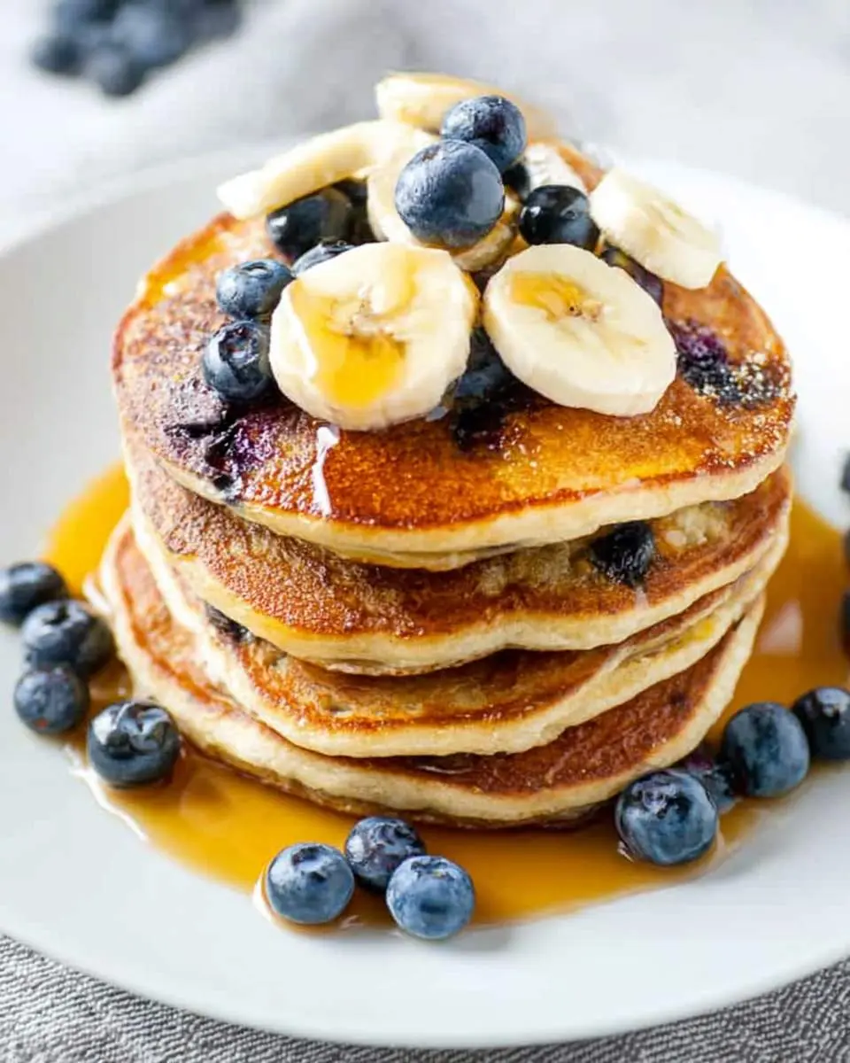 A delicious stack of golden Blueberry Banana Pancakes topped with fresh blueberries, banana slices, and maple syrup.
