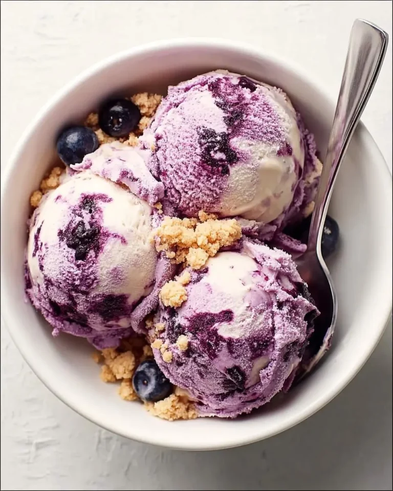 Three scoops of creamy Blueberry Cheesecake Ice Cream with cookie crumbles and fresh blueberries in a white bowl with a spoon.