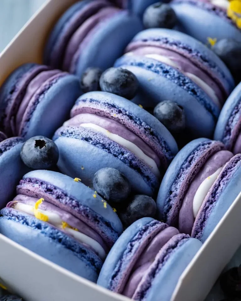 A white box filled with vibrant blue and purple Blueberry Lemonade Macarons, garnished with fresh blueberries and yellow lemon zest.
