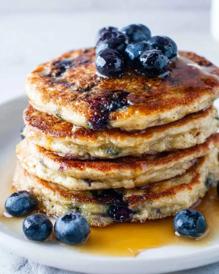 A delicious stack of golden-brown Blueberry Pancakes generously topped with fresh blueberries and drizzled with maple syrup.