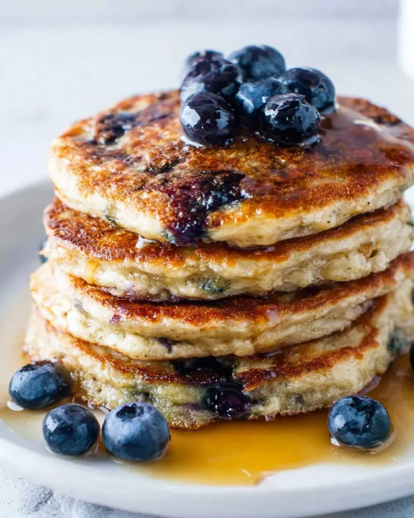 A delicious stack of golden-brown Blueberry Pancakes generously topped with fresh blueberries and drizzled with maple syrup.