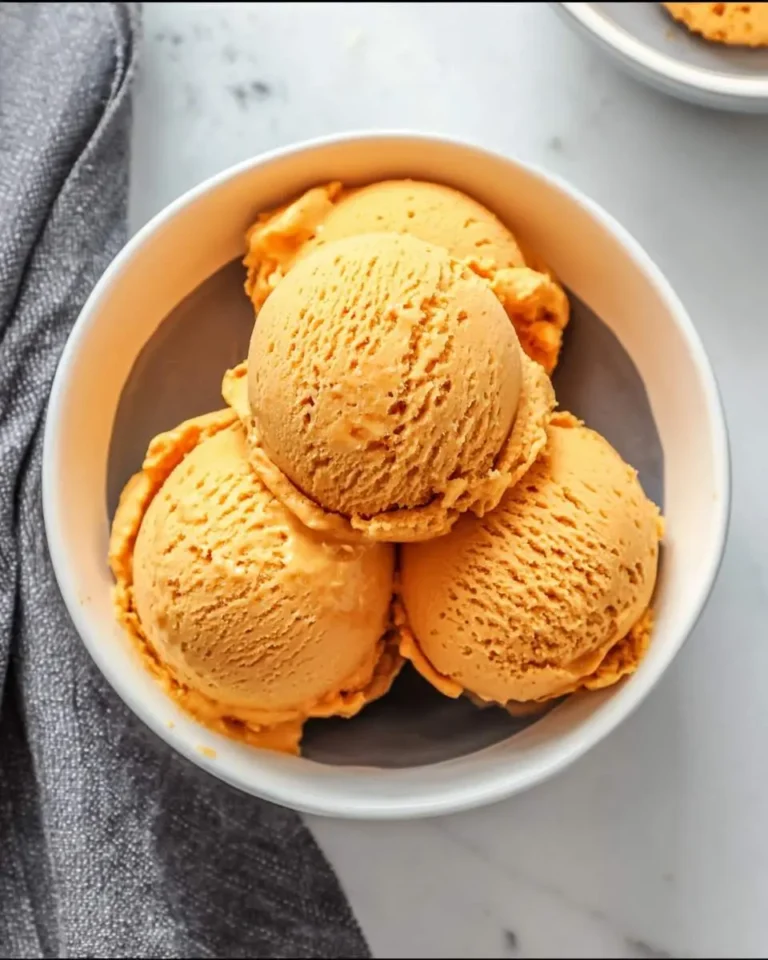 A white bowl filled with three scoops of rich orange Carrot Ice Cream on a white marble counter with a grey napkin.