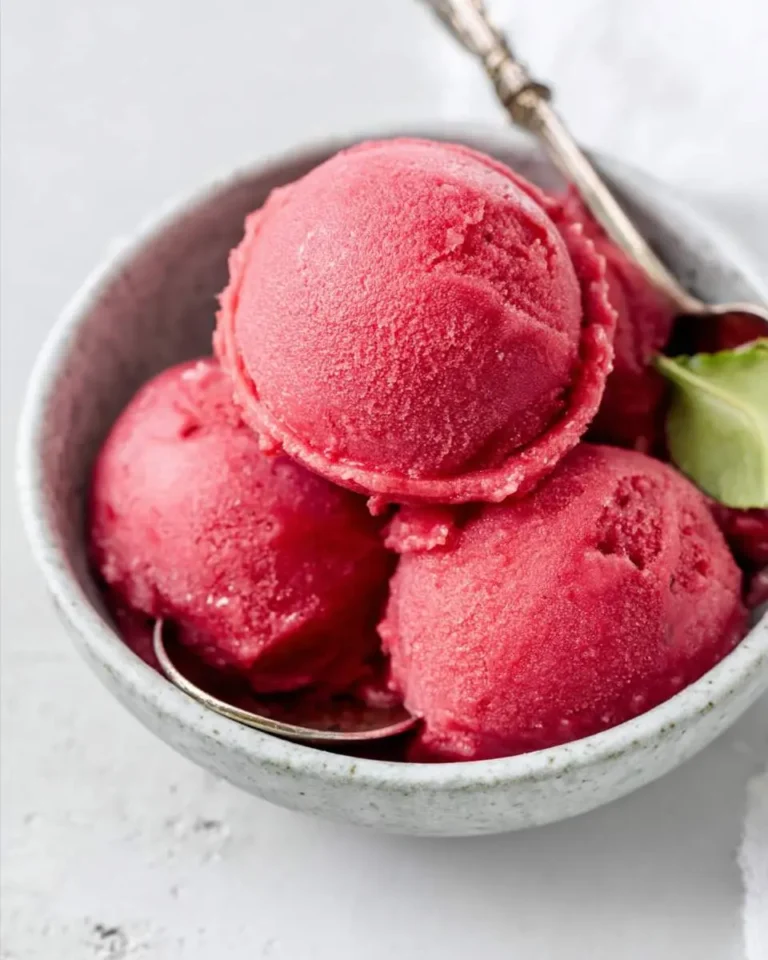 Several scoops of vibrant red cherry sorbet in a speckled bowl with a silver spoon and a green leaf garnish.