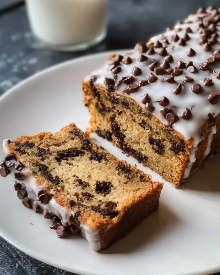 A freshly baked chocolate chip cookie bread loaf, glazed and topped with mini chocolate chips, with a slice on a white plate.