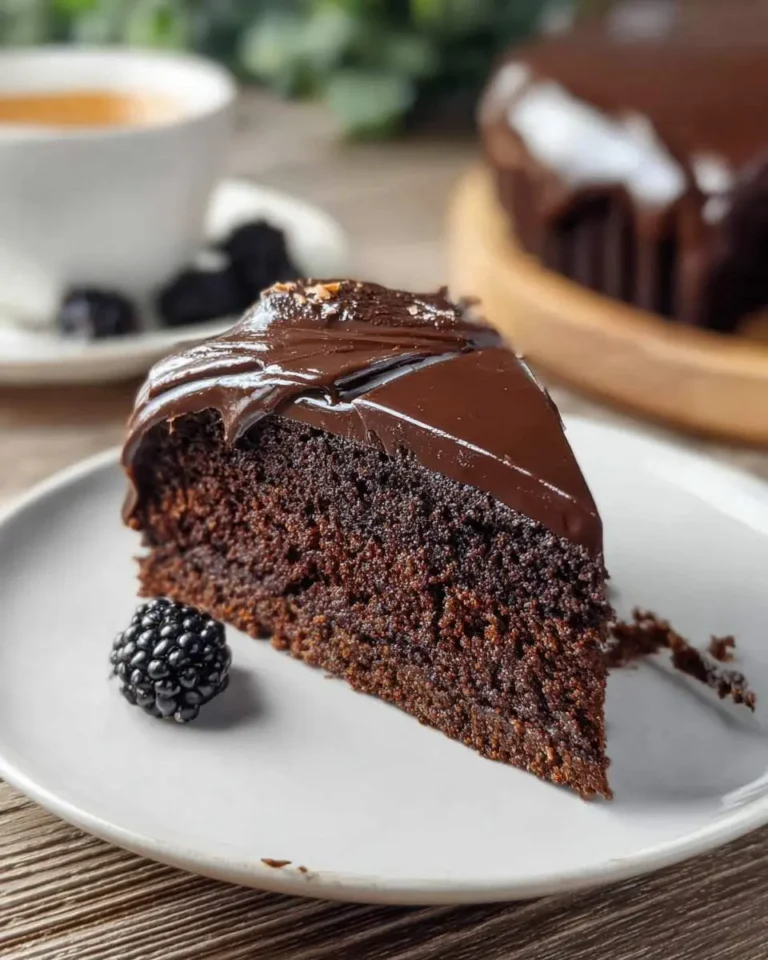 A slice of rich chocolate ganache cake with glossy frosting on a white plate, served with a blackberry.