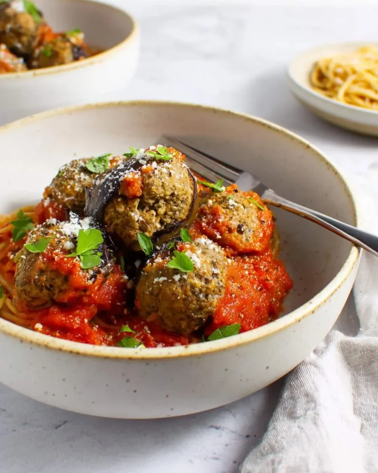 A close-up of savory eggplant meatballs with rich tomato sauce served over spaghetti in a speckled ceramic bowl, garnished with fresh herbs and grated cheese.