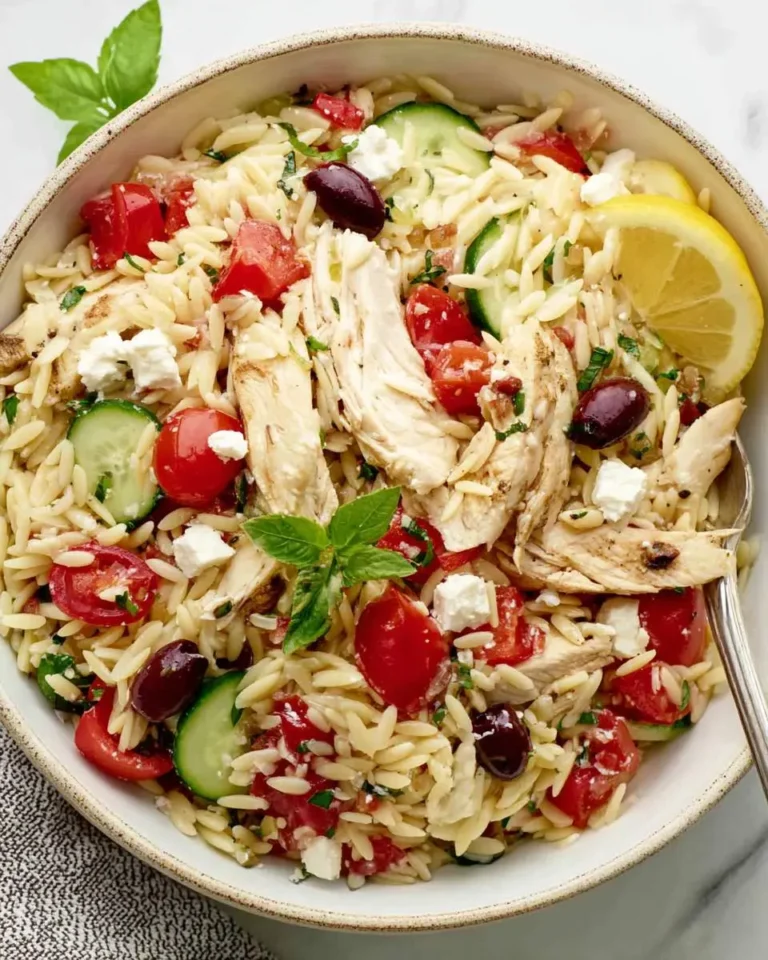A vibrant Greek Chicken Orzo Bowl with tender chicken, tomatoes, cucumber, feta, and olives, garnished with fresh basil and lemon.