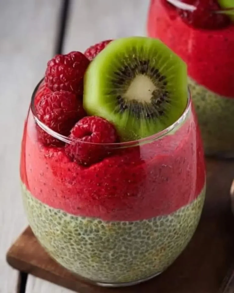 A close-up of a delicious Kiwi Raspberry Chia Pudding in a glass, with green chia seeds, red fruit puree, topped with fresh raspberries and a kiwi slice.