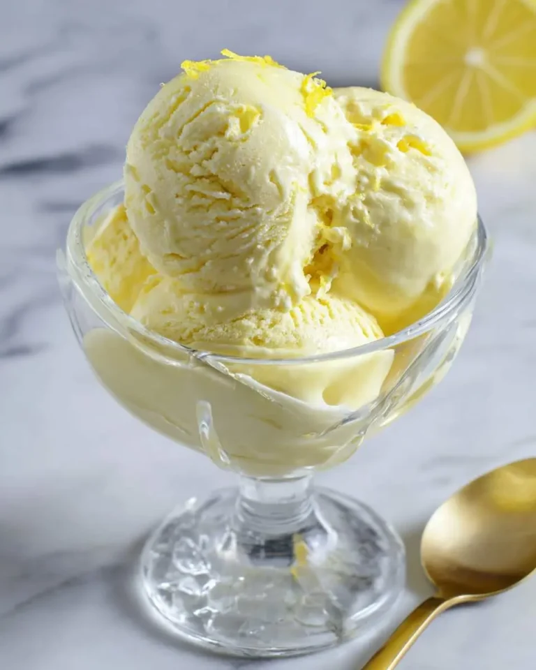 Three scoops of creamy lemon custard ice cream with lemon zest in a glass dish on a marble surface with a golden spoon.
