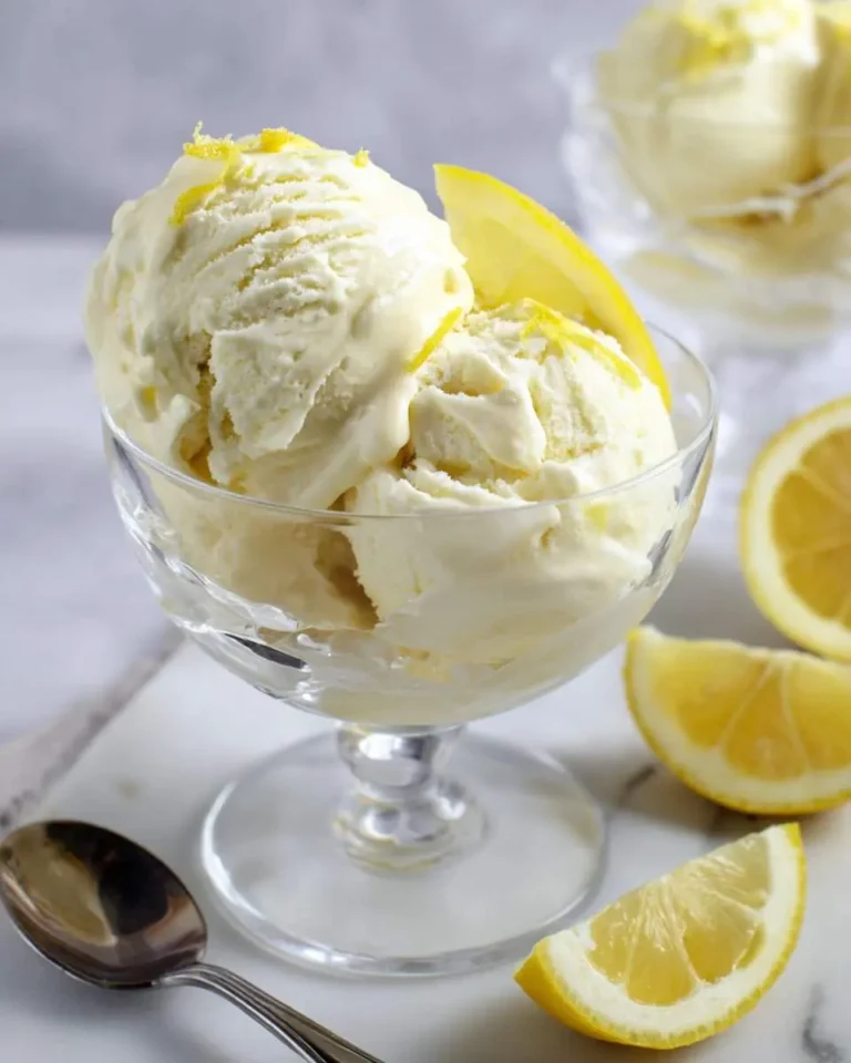 Three scoops of creamy lemon custard ice cream topped with zest and a lemon slice in a glass dessert cup with lemon wedges.