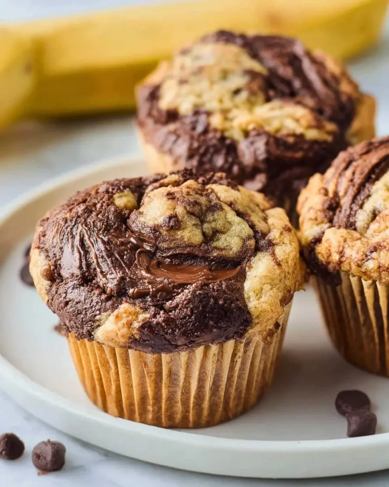 Three Nutella Banana Swirl Muffins on a white plate, showcasing a rich chocolate swirl and melted chocolate, with a banana blurred behind.