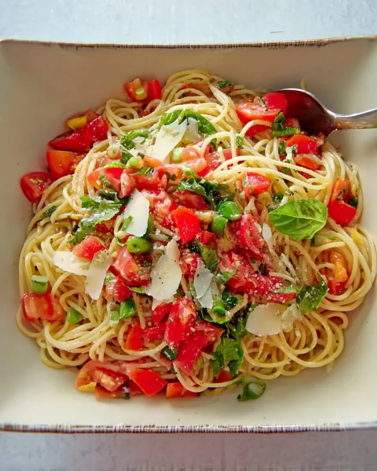 A close-up of a bowl of fresh Pasta alla Checca featuring spaghetti with bright red chopped tomatoes, green basil, and shaved Parmesan cheese, with a spoon in it.
