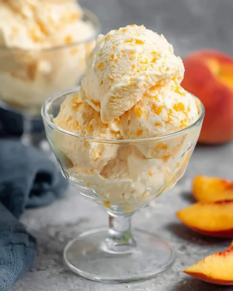Three scoops of creamy peach ice cream with visible peach chunks served in a clear glass dessert bowl, with fresh peaches nearby.