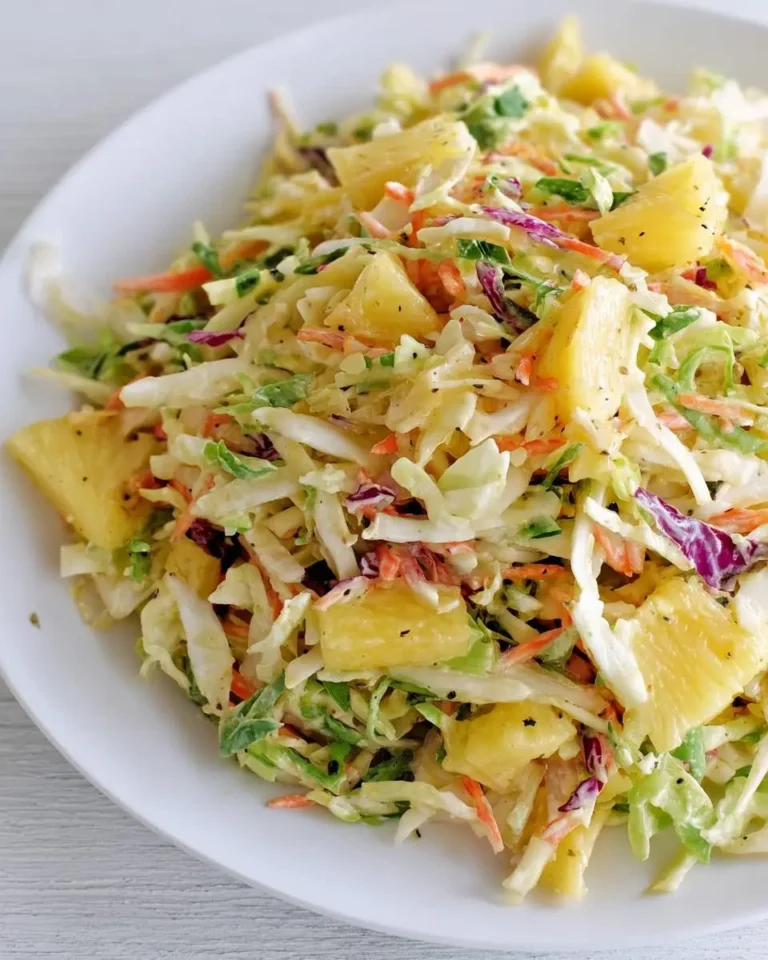 A close-up shot of a white plate piled high with vibrant pineapple coleslaw, featuring shredded cabbage, carrots, and large chunks of pineapple.