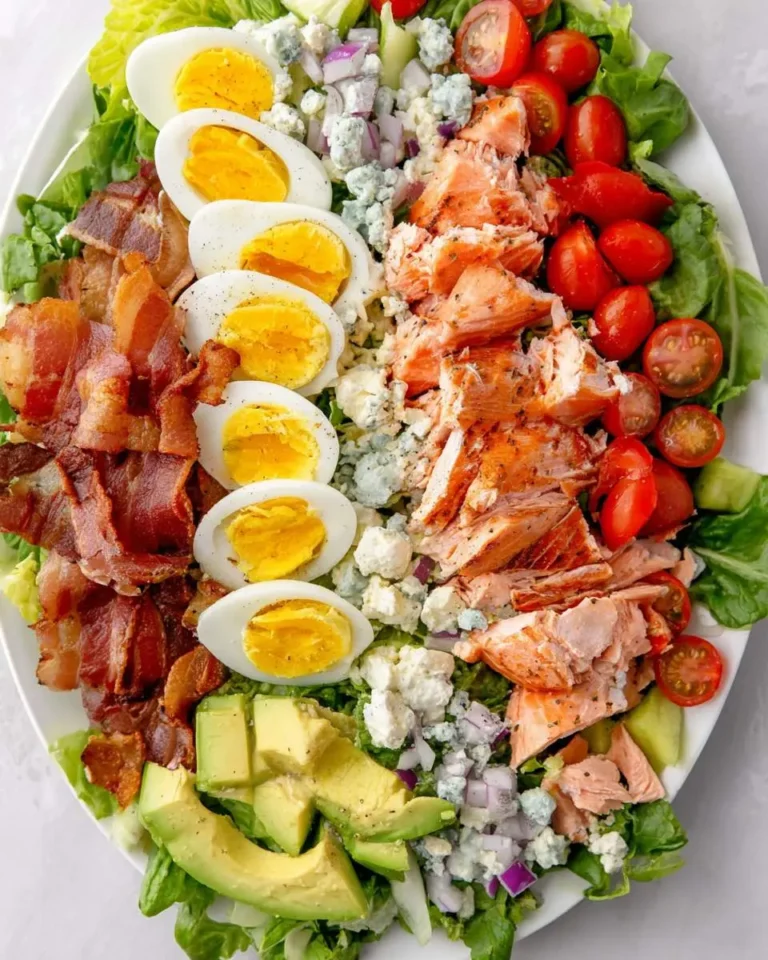 Overhead view of a Salmon Cobb Salad with eggs, bacon, avocado, tomatoes, and blue cheese on a white platter.