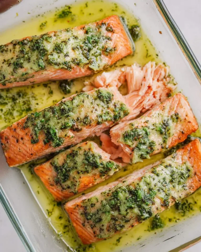 Close-up of cooked Salmon with pesto butter in a clear baking dish, with one fillet flaked and ready to serve.