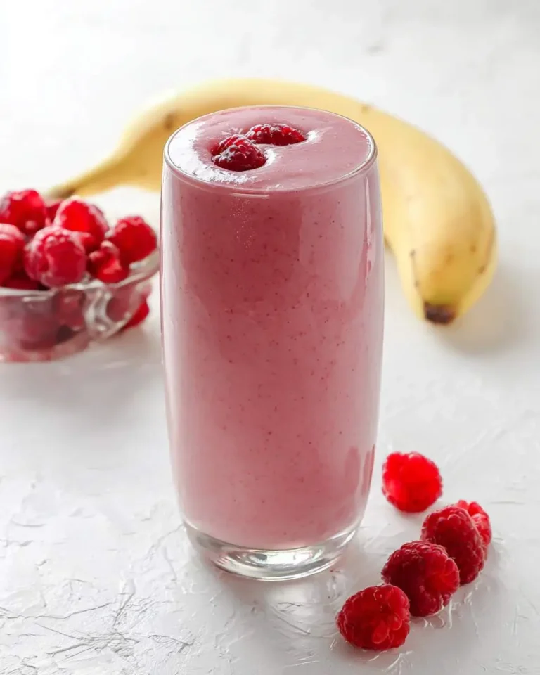 A tall glass of creamy banana raspberry smoothie topped with fresh raspberries, with a banana and berries in the background.