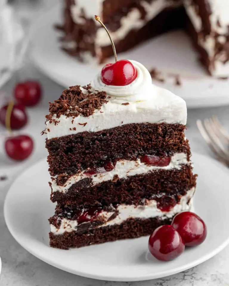 A slice of layered Black Forest Cake topped with whipped cream and a bright red cherry on a white plate.
