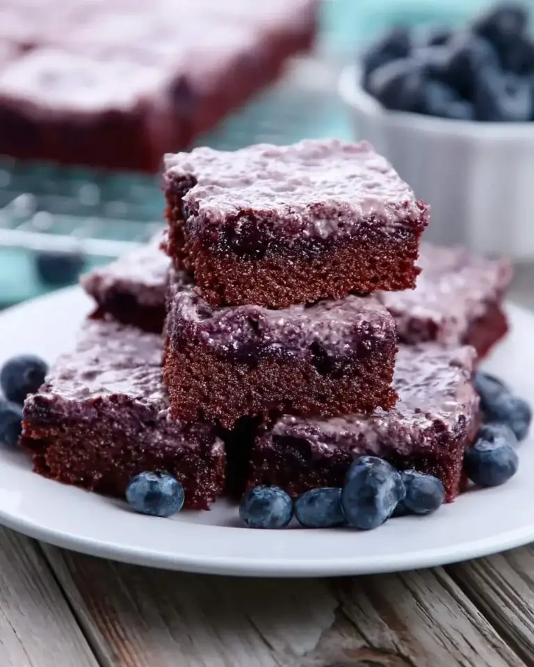 Stack of moist blueberry brownies with purple glaze on a white plate, surrounded by fresh blueberries.